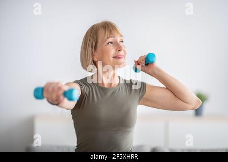 Inländisches Sporttraining. Positive ältere Frau macht Übungen mit Kurzhanteln und stärkt ihren Körper zu Hause Stockfoto