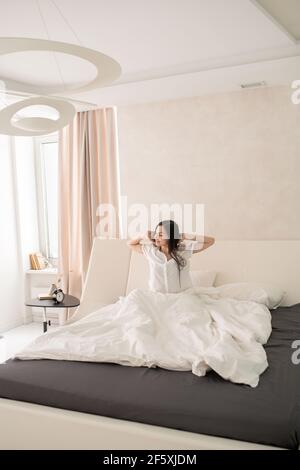 Glücklich junge schöne Brünette Frau Stretching, während auf großen sitzen Bequemes Doppelbett unter weißer Decke am Morgen Stockfoto