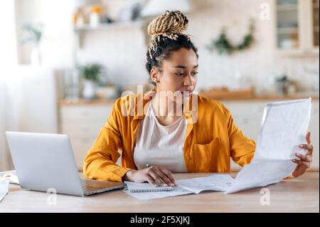 Konzentrierte, clevere afroamerikanische Frau, Studentin oder Designerin, in stilvoller Freizeitkleidung, in der Ferne studieren oder arbeiten, mit einem Laptop arbeiten, Blaupausen ansehen und untersuchen. Arbeiten Sie von zu Hause aus Stockfoto