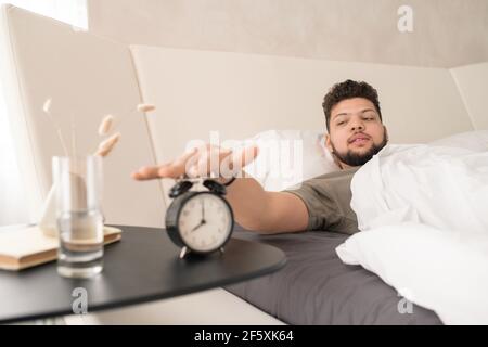 Junger bärtiger Mann im T-Shirt, der den Wecker drückt Liegen unter weißer Decke im großen Bett nach dem Schlafen Am Morgen Stockfoto