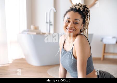 Süßes, athletisches afroamerikanisches Brünette Mädchen mit Dreadlocks, in Sportswear, auf dem Boden auf einer Fitnessmatte sitzend, vor dem Heimsport-Workout ein Warm-up machst, auf die Kamera schauend und fröhlich lächelnd Stockfoto