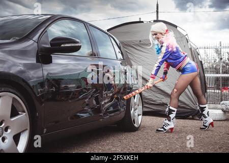 Moskau, Russland - 06. Juli 2020: Harley Quinn trifft mit einem Baseballschläger auf ein geparktes schwarzes Auto. Es ist eine Delle im Auto, das Auto ist beschädigt. Cosplay, Mädchen verkleidet als Charakter Kostüm Stockfoto