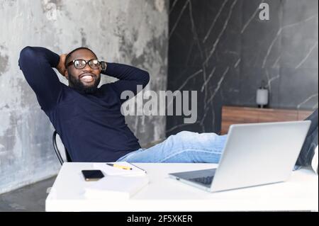 Glücklicher reifer afroamerikanischer Mann, der am Arbeitsplatz mit Laptop und Füßen auf dem Tisch sitzt und seine Hände hinter dem Kopf hält. Junge IT-Spezialisten machen eine Pause von der Arbeit. Entspanntes Arbeiten in geschlossenen Räumen Stockfoto