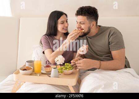Junge fröhliche Frau, die Sandwich mit Marmelade in ihren Mann Mund, während beide im Doppelbett sitzen und das Frühstück genießen Am Morgen Stockfoto