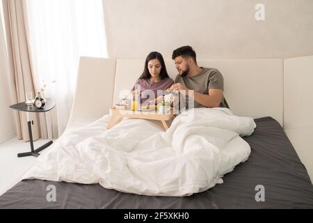 Junge liebevolle Paar in T-Shirts im Bett sitzen und mit Leckeres Frühstück, während weibliche Ausbreitung Marmelade auf Scheibe Weizen Brot Stockfoto
