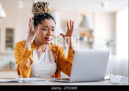 Angespannte Empörung frustriertes afroamerikanisches Mädchen mit Dreadlocks, Geschäftsfrau, Managerin oder Freiberuflerin, die aus der Ferne an einem Laptop arbeitete, Stress bei der Arbeit erlebte, schlechte Botschaft erhielt, mit Händen gestikulierte Stockfoto