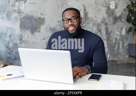 Porträt eines ernsten tausendjährigen afroamerikanischen Geschäftsmannes, der eine Brille trägt und die Kamera anschaut, am weißen Schreibtisch sitzt, mit einem Laptop arbeitet, selbstbewusster männlicher Arbeiter oder Regisseur, der im modernen Büro posiert Stockfoto