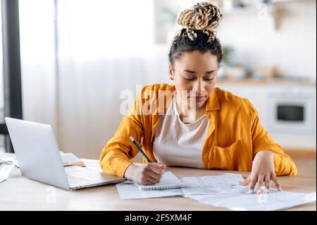 Selbstbewusste, intelligent fokussierte afroamerikanische Studentin oder Designerin, die stilvolle Freizeitkleidung trägt, fernab studiert oder arbeitet, Laptop benutzt, Notizen macht, Blaupausen untersucht. Arbeiten Sie von zu Hause aus Stockfoto