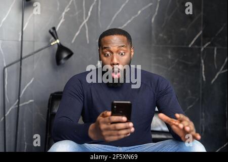 Erstaunt glücklich biracial Mann hält Telefon, Blick auf Handy, lesen gute Nachrichten in sms, sitzen auf Sessel, erstaunt schwarzen Kerl Gewinner begeistert von mobilen App, Sieg schreien vor Freude, feiern seinen Sieg Stockfoto