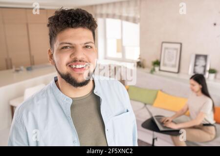 Happy junge bärtige Mann in Casualwear Blick auf Sie mit Toothy Lächeln gegen seine Frau auf der Couch sitzen und mit Laptop im Wohnzimmer Stockfoto