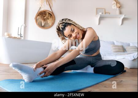 Flexible junge, schöne afroamerikanische Frau in Sportkleidung und mit Dreadlocks, engagiert sich für Fitness, führt einen gesunden Lebensstil, dehnt sich zu Hause auf einer Fitnessmatte, schaut auf die Kamera, lächelt Stockfoto