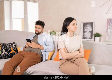Junge unzufriedene Frau in Casualwear überqueren Arme nach Brust, während Sitzend auf der Couch gegen ihren Mann scrollend im Smartphone Stockfoto