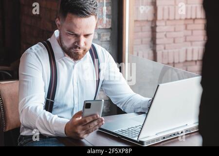 Junger Geschäftsmann, der auf sein Telefon schaut. Laptop auf dem Tisch vor ihm Stockfoto