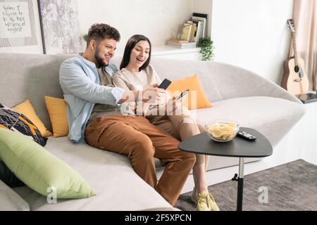 Glückliche junge Mann und Frau in Casualwear Blick auf ihre Smartphones werden angezeigt, während sie auf der Couch nahe beieinander sitzen Vor dem fernseher Stockfoto