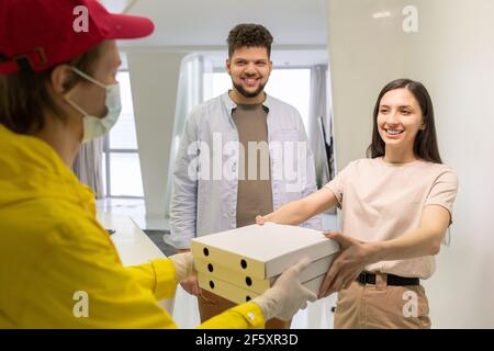 Glücklich junge Brünette weiblich in Casualwear suchen mit toothy Lächeln Beim männlichen Kurier, der ihren Stapel der Kisten mit bestellt vorbeigeht Pizza Stockfoto