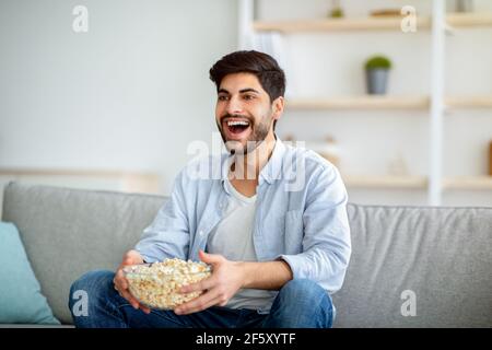Fröhlicher arabischer Mann, der Fernsehen schaut und Popcorn isst und sich auf der bequemen Couch im Wohnzimmer entspannt Stockfoto