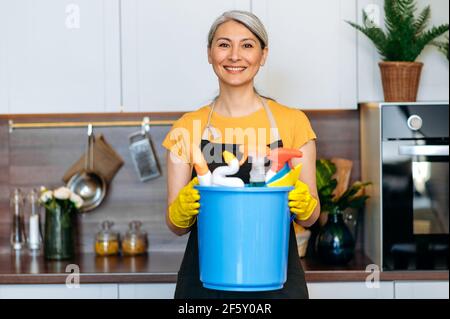 Schöne ältere grauhaarige asiatische Hausfrau oder Putzfrau in einer Schürze und Handschuhe stehen in der Küche, hält blauen Eimer mit Lumpen und Haushaltschemikalien, schaut auf die Kamera, lächelt Stockfoto