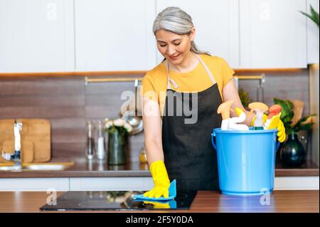 Schöne ältere grauhaarige asiatische Putzfrau oder Hausfrau in einer Schürze und Handschuhe steht in der Küche, wischen den Herd, mit einem Eimer Reinigungsmittel, lächelnd. Reinigungsservice Stockfoto