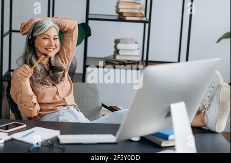 Selbstbewusst entspannt schöne ältere grauhaarige asiatische Frau, Geschäftsführerin, Manager, freiberuflich, im Gespräch mit Kollegen oder Freunden per Videoanruf, warf ihre Beine auf den Tisch, lächelte freundlich Stockfoto