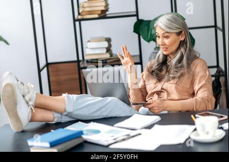 Attraktive erfolgreiche Senior grauhaarige asiatische Frau, Manager, Business Leader, Immobilienmakler, im Gespräch mit Freunden, Kollegen oder Familie per Videoanruf, ihre Füße auf den Tisch werfen, winkt mit der Hand Stockfoto