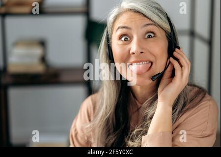 Porträt einer glücklichen törichten, reifen grauhaarigen asiatischen Frau im Headset, Callcenter-Mitarbeiter, Manager, Freiberufler, Blick auf die Kamera, Herumtäuschen, Zunge zeigen, Spaß haben, lustiges Gesicht Stockfoto