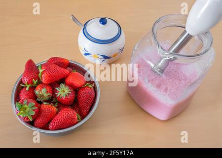 Schüssel voller Erdbeeren und einen Krug mit frisch gemahlener Erdbeermilchshake, eine Zuckerschüssel mit einem Löffel darin, einen Stabmixer im Krug Stockfoto