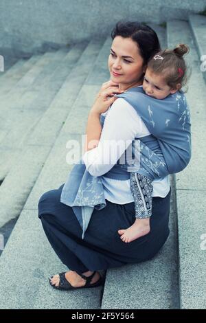Mutter mit Baby Umhüllung in grauem Tragetuch Rücken sitzen auf Granittreppen Stockfoto