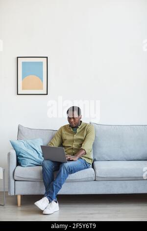 Vertikales Porträt eines erwachsenen afroamerikanischen Mannes mit Laptop, während er auf der Couch in minimalem Innenraum sitzt, Kopierer Platz Stockfoto