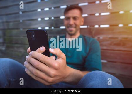 Junger lächelnder Mann mit seinem schwarzen Smartphone im Freien bei Sonnenuntergang. Smartphone betrachten. Mann mit einem facetime Videoanruf Stockfoto
