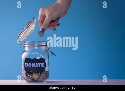 Eine Münze in ein Glasgefäß mit Münzen mit einem Kreide-Spendenschild auf blauem Hintergrund legen. Spenden- und Wohltätigkeitskonzept Stockfoto