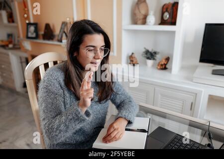 Medium shot junge taube Frau trägt eine Brille und ein Pullover ist mit Gebärdensprache Namen buchstabieren, während in einem Online-Videoanruf in ihrem Wohnzimmer. Stockfoto