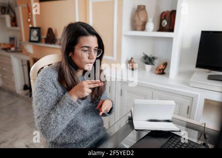 Medium shot junge taube Frau trägt eine Brille und ein Pullover ist mit Gebärdensprache Namen buchstabieren, während in einem Online-Videoanruf in ihrem Wohnzimmer. Stockfoto