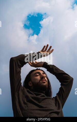 Porträt eines jungen Mannes mit erhobenen Armen. Tragen einen grünen Rollkragenpullover, entspannt in seinen Gedanken. Meditierender, langhaariger Mann, der sich mit seinem verbindet Stockfoto