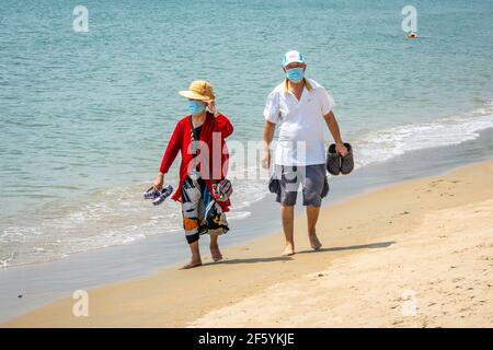 Sanya China , 24 März 2021 : Chinesisches Paar von älteren Menschen zu Fuß barfuß auf Sanya Bucht Strand tragen blaue chirurgische Gesichtsmasken, um Covid zu verhindern Stockfoto