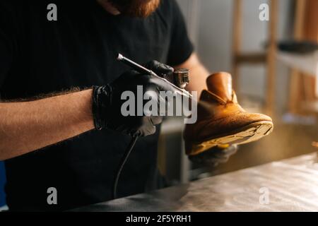 Nahaufnahme Hände von nicht erkennbaren Schuster tragen schwarze Handschuhe Sprühen Farbe von hellbraunen Lederschuhe. Stockfoto