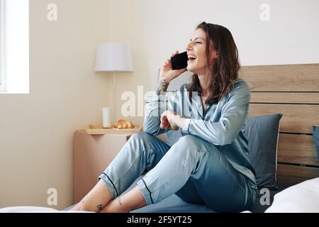 Aufgeregt schöne junge Frau im Schlafanzug sitzen auf dem Bett und Telefonieren mit einem Freund Stockfoto
