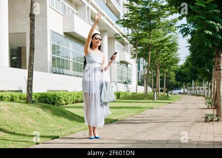 Hübsche junge Frau in Sonnenbrille hält Smartphone und winkt mit Hand, um ein Taxi zu bekommen Stockfoto