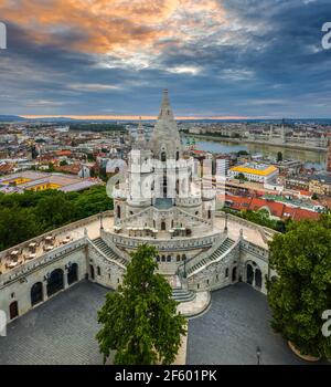 Budapest, Ungarn - Luftaufnahme der berühmten Fischerbastei bei Sonnenaufgang mit bunten Wolken und Parlamentsgebäude im Hintergrund. Grüne Bäume, Stockfoto