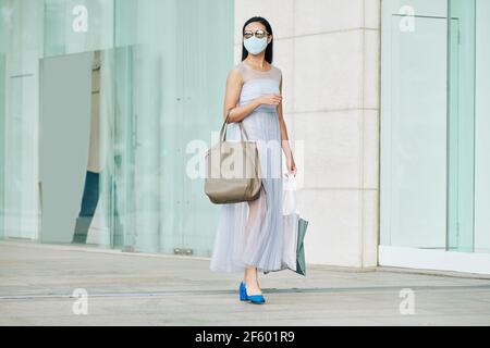 Schöne stilvolle junge Frau in Sonnenbrille und medizinische Maske zu Fuß Auf der Straße mit Papiertüten nach dem Einkaufen Stockfoto