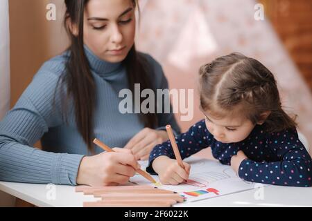 Adirable kleines Mädchen mit ihrer schönen jungen Mutter zeichnet in das Album mit Farbe Pensins. Nettes Mädchen malt Malvorlagen zu Hause während quararntin Stockfoto