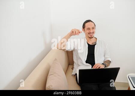 Glücklich lächelnder Mischling Mann sitzt auf dem Sofa zu Hause und Arbeiten mit einem Laptop Stockfoto