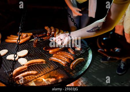 Machen Grill in der Nacht. Menschliche Hand, die gegrillte Wurst vom Grill im Freien nimmt. Open Air Camp Stockfoto