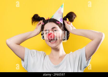 April ist ein Narrentag. Porträt einer fröhlichen Frau in einer Mütze und mit einer roten runden falschen Nase, writthing lustig. Gelber Hintergrund. Stockfoto