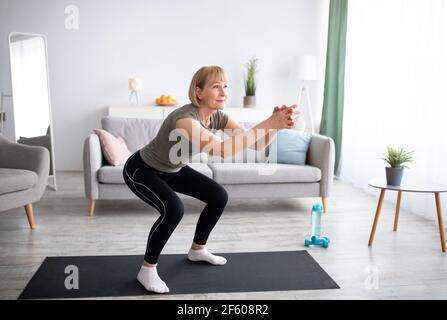 Positive ältere Dame macht Kniebeugen auf häusliche Training im Wohnzimmer, freien Platz Stockfoto