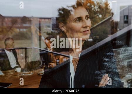 Geschäftsfrau schaut durch das Fenster, während männliche Kollegen im Büro diskutieren Im Hintergrund Stockfoto