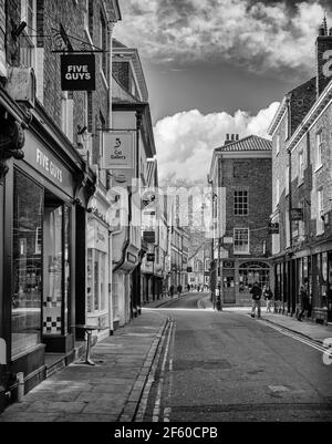 Eine alte Straße in York. Alte Gebäude befinden sich auf jeder Seite und Sonnenlicht erstreckt sich über eine Kreuzung. Geschäfte säumen die Straße und ein wolkiger Himmel ist oben. Stockfoto