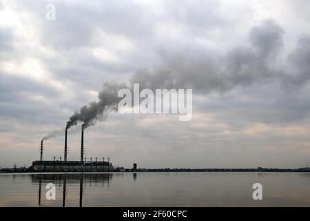 Kohlekraftwerk hohe Rohre mit schwarzem Rauch, der nach oben die verschmutzende Atmosphäre mit den Reflexionen von ihm im Seewasser bewegt. Stockfoto