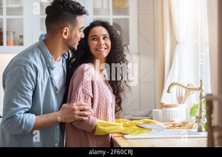 Liebevoller Arabischer Ehemann Umarmt Frau Aus Dem Rücken, Während Sie Geschirr Waschen Stockfoto