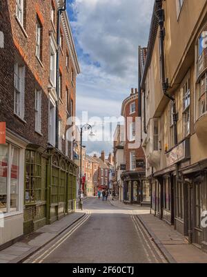 Eine alte Straße in York. Alte Gebäude befinden sich auf jeder Seite und Sonnenlicht erstreckt sich über eine Kreuzung. Geschäfte säumen die Straße und ein wolkiger Himmel ist oben. Stockfoto