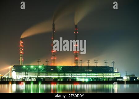 Hell erleuchtetes Kohlekraftwerk hohe Rohre mit schwarzem Rauch, der nach oben die verschmutzende Atmosphäre in der Nacht mit den Reflexen von ihm im Seewasser bewegt. Stockfoto